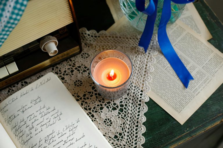 image of a desktop containing a notebook with written words, a lit candle, a lace doily and pages from a book.